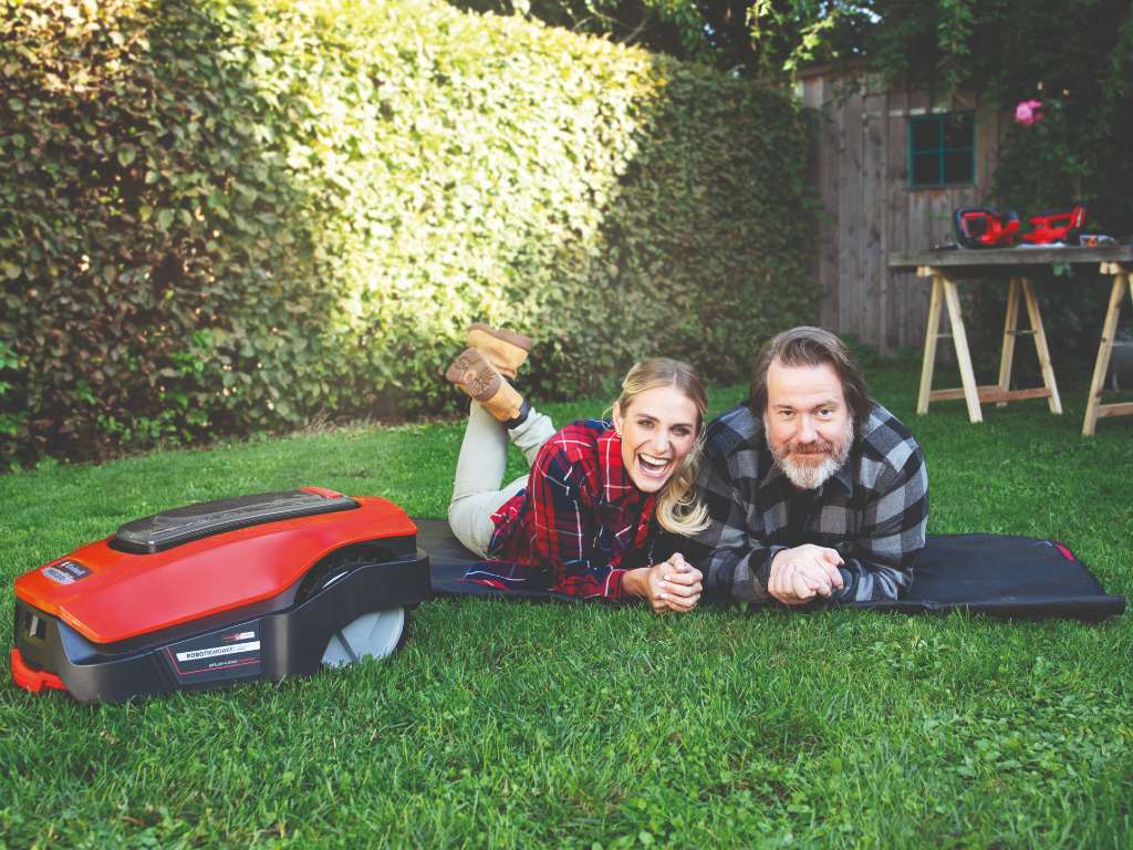 A man and a woman lie in the grass next to a robotic lawnmower