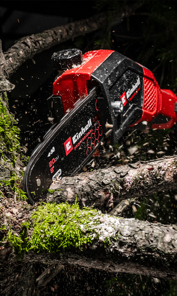 A red Einhell cordless chainsaw cuts through a branch with sawdust flying through the air.